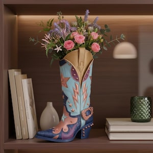 Op de afbeelding: Een decoratieve cowboylaarsvaas in tinten blauw, goud en paars, gevuld met roze rozen en andere bloemen. De laars heeft ster- en bliksemontwerpen, een gesp en een bezaaide hak. De vaas staat op een plank met boeken en een groene glazen container.