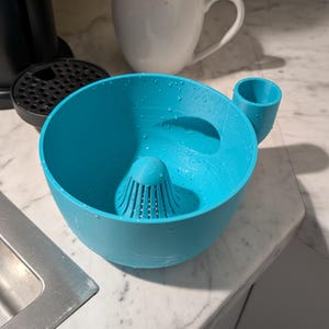 Op de afbeelding: Een helder turquoise citruspers met een ingebouwde zeef en een klein kopje. De citruspers staat op een wit marmeren aanrecht. Een witte mok met een blauwe rand staat op de achtergrond.