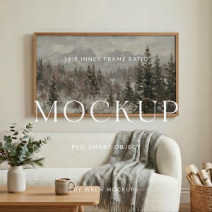 Puede incluir: Una pintura de paisaje enmarcada con una escena de montaña y bosque cuelga sobre un sofá blanco. La pintura está en un marco de madera marrón claro. Una manta gris está sobre el sofá. Una mesa de centro y un jarrón de madera están en primer plano.