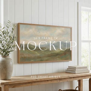 Op de afbeelding: Een ingelijst landschapsschilderij met een lichtbruine houten lijst hangt aan een witte muur. Het schilderij toont een berglandschap in gedempte groene, blauwe en bruine tinten. De tekst "16:9 FRAME TV MOCKUP" staat over de afbeelding.