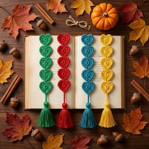 Puede incluir: Cuatro marcapáginas de ganchillo con forma de corazón en verde, rojo, azul y amarillo. Cada marcapáginas presenta una cadena vertical de cinco corazones que termina en una borla a juego, sobre las páginas abiertas de un libro.