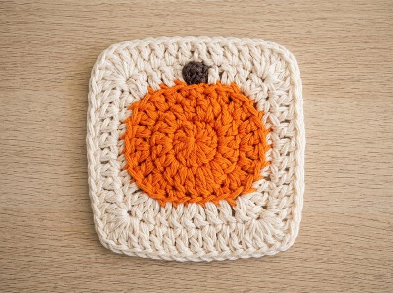May include: A square crochet coaster featuring a circular orange pumpkin motif centred within a cream-coloured border. The pumpkin is worked in a vibrant orange yarn with a small, dark brown stem at the top, all crafted with visible crochet stitches.