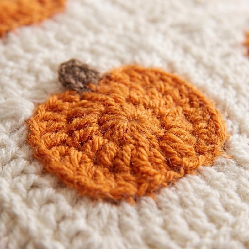 May include: A textured crochet fabric featuring a repeating pattern of small, orange pumpkins with brown stems set against a cream-coloured background. The yarn work displays a distinct, raised stitch construction that creates a tactile surface.
