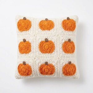 May include: A square crochet throw pillow featuring a grid of nine off-white squares, each containing a textured orange pumpkin motif with a small brown stem. The pillow is constructed with a visible yarn stitch pattern throughout the entire surface.