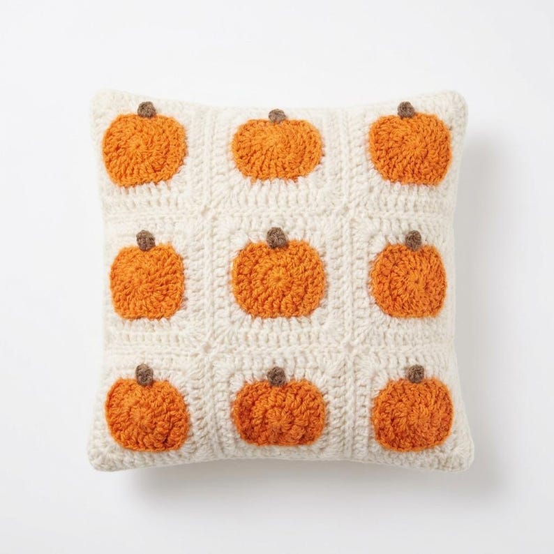 May include: A square crochet throw pillow featuring a grid of nine off-white squares, each containing a textured orange pumpkin motif with a small brown stem. The pillow is constructed with a visible yarn stitch pattern throughout the entire surface.