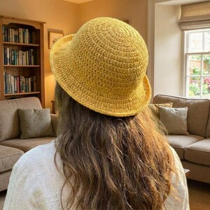 May include: A yellow crochet hat with a wide brim. The hat is made of a textured yarn and has a rounded crown. The hat is worn on a person's head, with long brown hair visible. The background includes a bookshelf and a sofa.