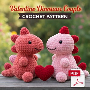 May include: Two crocheted dinosaur plushies, one red and one pink, sit side-by-side with a red heart. The text "Valentine Dinosaur Couple CROCHET PATTERN" is at the top. A PDF icon is in the lower right corner.