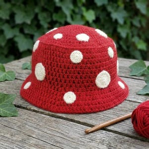 May include: A red crochet bucket hat featuring a pattern of circular white spots across the crown and brim. The hat is constructed with a dense, textured stitch pattern and is accompanied by a wooden crochet hook and a ball of red yarn.