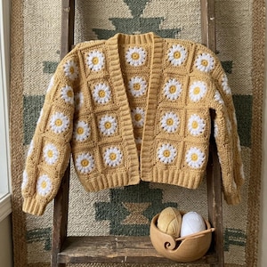 Puede incluir: Un cárdigan de crochet amarillo mostaza con cuadrados de margaritas blancas y amarillas. El suéter abierto tiene mangas largas y se exhibe en una escalera de madera. Un cuenco de madera con hilo y un ganchillo está en primer plano.