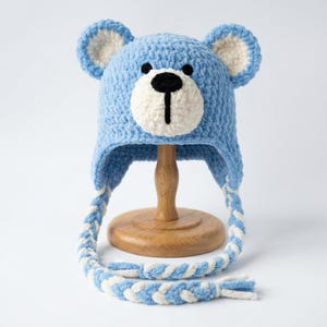May include: A light blue crochet hat featuring a bear face design with a white muzzle, black nose, and two rounded ears. The hat includes long, braided earflaps made of alternating light blue and white yarn strands, creating a soft texture.