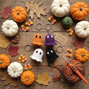 May include: Four crocheted ghost keychains in orange, purple, white, and black, each with two white eyes. A brown paper tag reads Handmade Cozy Ghost. A ball of orange and black yarn with a crochet hook and various pumpkins are arranged nearby.