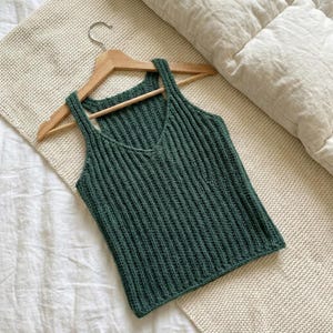 May include: A dark green, ribbed knit vest top with a V-neck and wide straps, displayed on a wooden hanger. The top is made of a textured material and has a simple, classic design. The vest top is approximately 51 cm long.