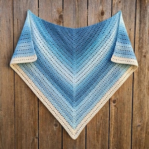 Pode incluir: Um xale triangular de crochê com um padrão de gradiente de cor que transita do creme claro nas bordas para vários tons de azul no centro. O tecido exibe um padrão de pontos repetitivo com fileiras horizontais e uma borda texturizada.