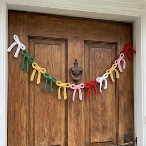 Puede incluir: Una colorida guirnalda de lazos de ganchillo en blanco, verde, amarillo, rosa y rojo cuelga de una puerta de madera. La guirnalda hecha a mano está ensartada, creando una decoración festiva. La puerta tiene un aldaba de latón.