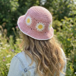 Puede incluir: Un sombrero de pescador rosa tejido a ganchillo con aplicaciones de margaritas blancas y amarillas. El sombrero tiene un borde blanco y está hecho de un hilo suave y texturizado. Un accesorio elegante para días soleados.
