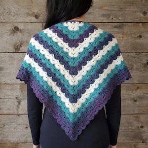 May include: A handmade, crocheted shawl in a triangular shape. The shawl features a scalloped edge and is striped with bands of white, teal, purple, and blue. The shawl is worn over a black top, with a wooden background.