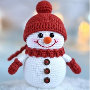 Könnte beinhalten: Gehäkelter Schneemann mit roter Mütze, Schal und Fäustlingen. Der Schneemann hat eine Karottennase, schwarze Augen und ein gesticktes Lächeln. Drei braune Knöpfe zieren die Vorderseite. Eine festliche Winterdekoration.