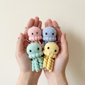 May include: Four colorful crochet jellyfish keychains in pink, blue, green, and yellow. Each jellyfish has black eyes and a silver keychain ring. The tentacles are long and flowing. The keychains are held in cupped hands.