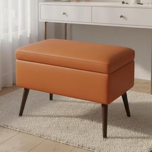 Peut inclure: Un banc de rangement orange avec des pieds en bois foncé repose sur un tapis de couleur claire. Le banc est rectangulaire avec un dessus rembourré. Une coiffeuse blanche avec un miroir rond et un rideau blanc sont en arrière-plan.