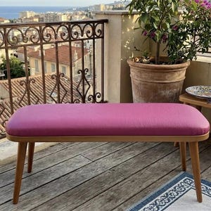 Puede incluir: Banco moderno con asiento magenta y patas de madera clara. El banco es rectangular con esquinas redondeadas y está colocado en una terraza de madera. El fondo presenta una barandilla de hierro forjado, un paisaje urbano y plantas en macetas.
