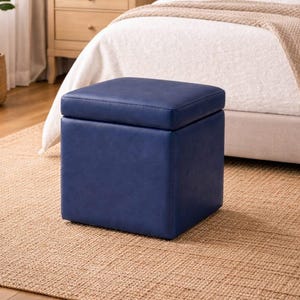 Könnte beinhalten: Ein marineblauer quadratischer Hocker mit einem hochklappbaren Deckel steht auf einem gewebten Jute-Teppich in einem Schlafzimmer. Der Hocker besteht aus einem glatten, einfarbigen Material und steht in der Nähe eines Bettes mit neutralfarbener Bettwäsche.