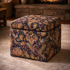 May include: A square ottoman with a hinged lid, upholstered in a paisley pattern. The fabric features a navy blue background with a colorful design of yellow, orange, and teal. The ottoman sits on a neutral-colored rug.