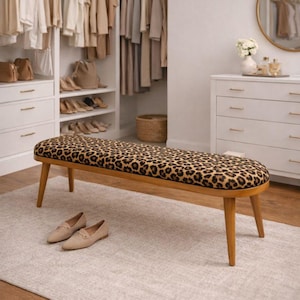 May include: A wooden bench with a leopard print seat cushion and four tapered legs. The bench is placed on a light gray rug in front of a white closet filled with clothing and shoes. A pair of beige loafers sits on the rug.