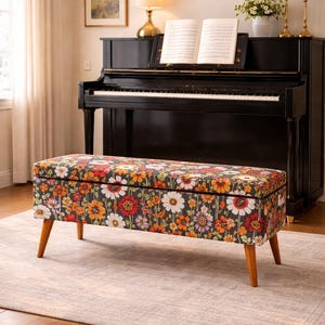 May include: A rectangular storage bench with a floral pattern in red, orange, and white on a dark background. The bench has wooden legs and is placed in front of a black piano. The room has a neutral color palette.