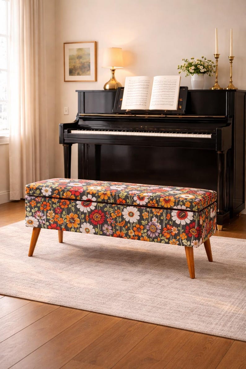 May include: A rectangular storage bench with a floral pattern in red, orange, and white on a dark background. The bench has wooden legs and is placed in front of a black piano. The room has a neutral color palette.