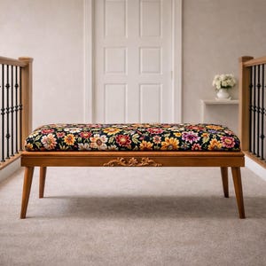 May include: Wooden bench with a floral cushion. The bench has four tapered legs and a decorative carved apron. The cushion features a black background with colourful flowers in shades of yellow, red, and purple. The bench is approximately 120 cm long.