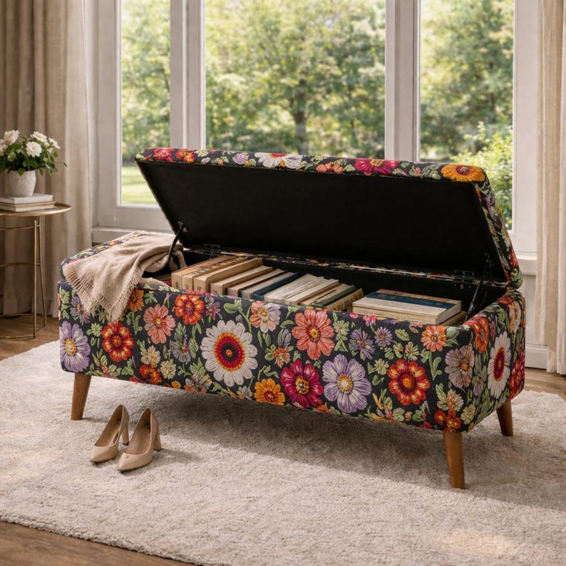 May include: A floral-patterned storage bench with a hinged lid, revealing books inside. The bench features a dark background with colorful flowers and wooden legs. A beige blanket rests on the bench, with a pair of beige heels in front.