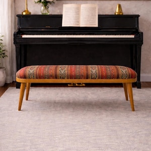 Op de afbeelding: Een bank in mid-century stijl met een stoffen zitting met patronen in tinten rood, oranje en bruin. De bank heeft een licht houten frame en vier schuine poten. De bank staat voor een zwarte piano.