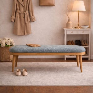 May include: A blue upholstered bench with wooden legs. A tan trench coat and a brown handbag hang on the wall. A lamp and side table are in the background, creating a cosy interior scene. The bench is approximately 120 cm long.