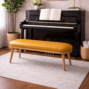 Puede incluir: Un banco amarillo mostaza con patas de madera se encuentra sobre una alfombra gris claro frente a un piano negro. Una planta en maceta y una cesta están a ambos lados del banco. El piano tiene partituras en el atril.