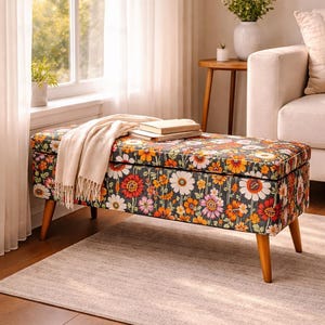 May include: Floral patterned storage bench with wooden legs. The bench features a dark background with colorful flowers in shades of orange, pink, and white. A beige throw blanket and books rest on top.