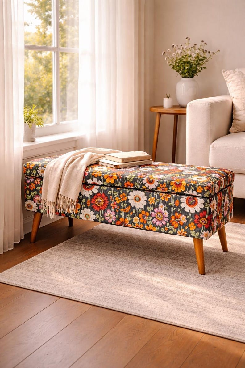 May include: Floral patterned storage bench with wooden legs. The bench features a dark background with colorful flowers in shades of orange, pink, and white. A beige throw blanket and books rest on top.