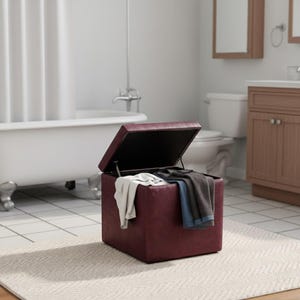 May include: A burgundy storage ottoman with a hinged lid, open to reveal folded towels and clothing. The square ottoman sits on a patterned rug in a bathroom setting. The bathroom has a white bathtub and a wooden cabinet.