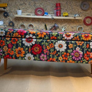 May include: A floral storage bench with wooden legs. The bench features a dark background with a vibrant pattern of orange, red, pink, and white flowers. The legs are a light brown color.