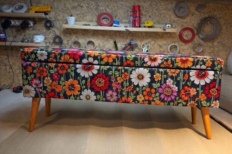 May include: A floral storage bench with wooden legs. The bench features a dark background with a vibrant pattern of orange, red, pink, and white flowers. The legs are a light brown color.