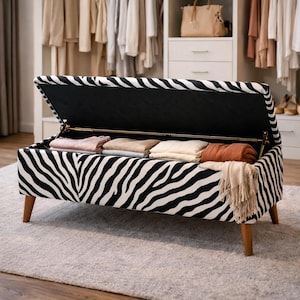 May include: A zebra-print storage bench with a hinged lid, revealing folded blankets and throws. The bench has wooden legs and a black interior. The overall design is a mix of black and white, creating a striking visual contrast.