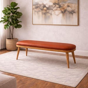 Może przedstawiać: Ławka w stylu mid-century z siedziskiem ze skóry w kolorze czerwonobrązowym i jasną drewnianą ramą. Ławka ma około 152 cm długości i cztery skośne nogi. Abstrakcyjny obraz wisi na ścianie za ławką.