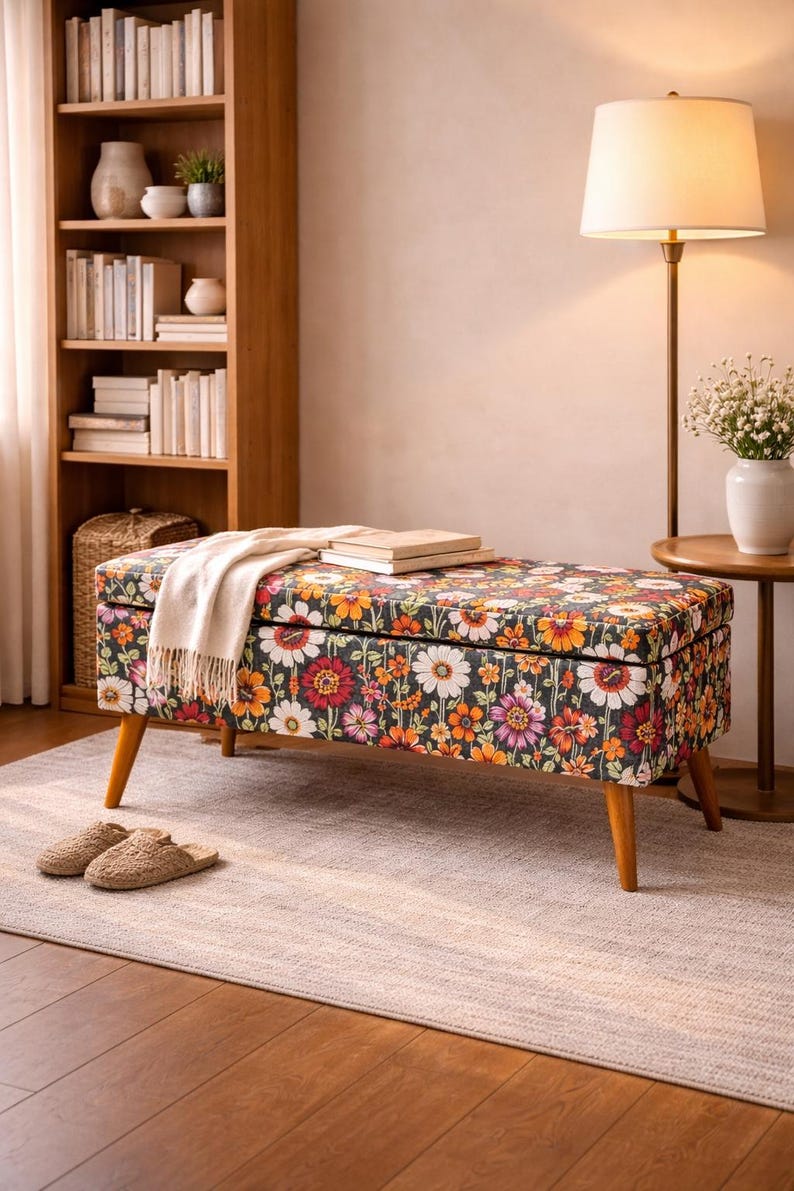 May include: A rectangular storage bench with a floral pattern in shades of orange, red, pink, and white on a dark background. The bench has wooden legs and is topped with a cream-colored throw and books. A pair of slippers sits on the rug.