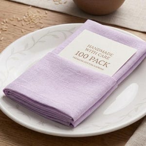 Może przedstawiać: Stos lawendowych serwetek z tkaniny na białym talerzu z kwiatowym wzorem. Karta z napisem "HANDMADE WITH CARE 100 PACK" jest wsunięta w serwetki. Po prawej stronie znajduje się widelec i gałązka lawendy.