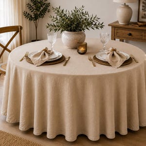 May include: A round beige tablecloth draped over a circular dining table. The table is set with two white plates, beige napkins, gold-toned forks and knives, two clear textured glasses, and a small glass candle holder with a lit candle inside.