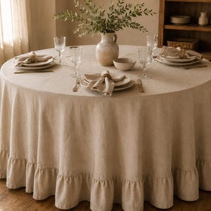 Può includere: Una tovaglia rotonda in lino beige con orlo a balze, disposta su un tavolo da pranzo con piatti bianchi, bicchieri trasparenti, tovaglioli di stoffa con anelli in legno, una piccola ciotola in ceramica e un vaso centrale con fogliame.