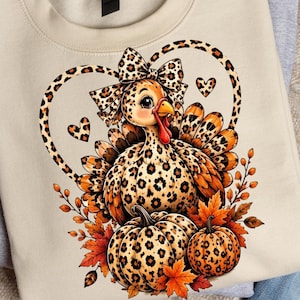 Puede incluir: Una sudadera beige de cuello redondo con un gráfico de un pavo con estampado de leopardo, que lleva un lazo a juego. El diseño incluye dos calabazas con estampado de leopardo, hojas de otoño naranjas y rojas, y corazones.