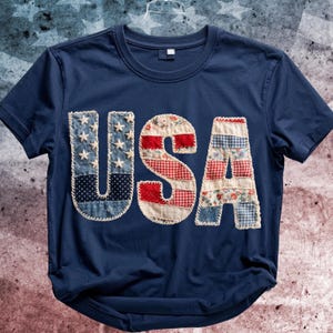 Puede incluir: Camiseta azul marino con la palabra "USA" aplicada en la parte delantera. Las letras están hechas de varias telas patrióticas, incluyendo mezclilla con estrellas blancas, cuadros rojos y estampados florales. Un diseño casual y festivo.