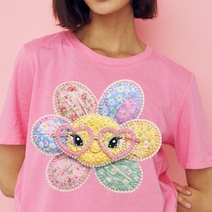 Op de afbeelding: Een roze t-shirt met een kleurrijk bloemdesign. De bloem heeft een gele kern, veelkleurige bloemblaadjes en een roze hartvormige bril. De bloemblaadjes hebben bloempatronen en zijn omrand met witte kralen.