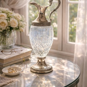 Può includere: Una brocca in argento e cristallo con manico e base decorativi. Il corpo in vetro trasparente presenta un motivo a taglio diamante. La brocca è appoggiata su un tavolo rotondo con piano in vetro. Un bouquet di rose bianche è sullo sfondo.