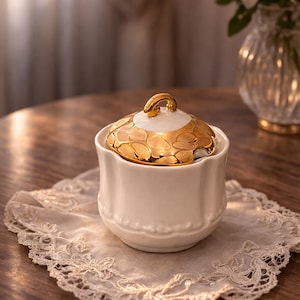 Könnte beinhalten: Eine weiße Zuckerdose aus Keramik mit einem goldfarbenen Deckel mit Blattmuster. Die Schale steht auf einer Spitzendecke, im Hintergrund eine Vase mit weißen Blumen. Der Griff am Deckel ist ebenfalls goldfarben.